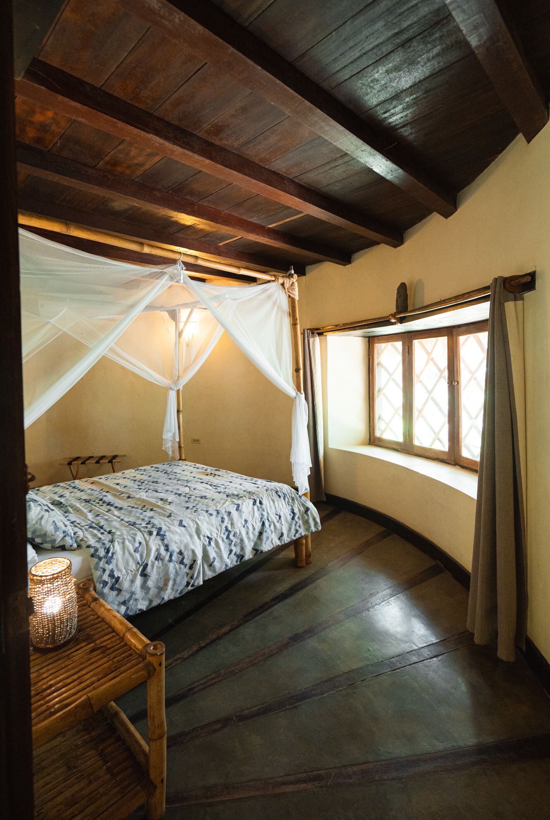 A room with a bed under a wooden ceiling, a window with curtains, and a bedside table with a lamp.