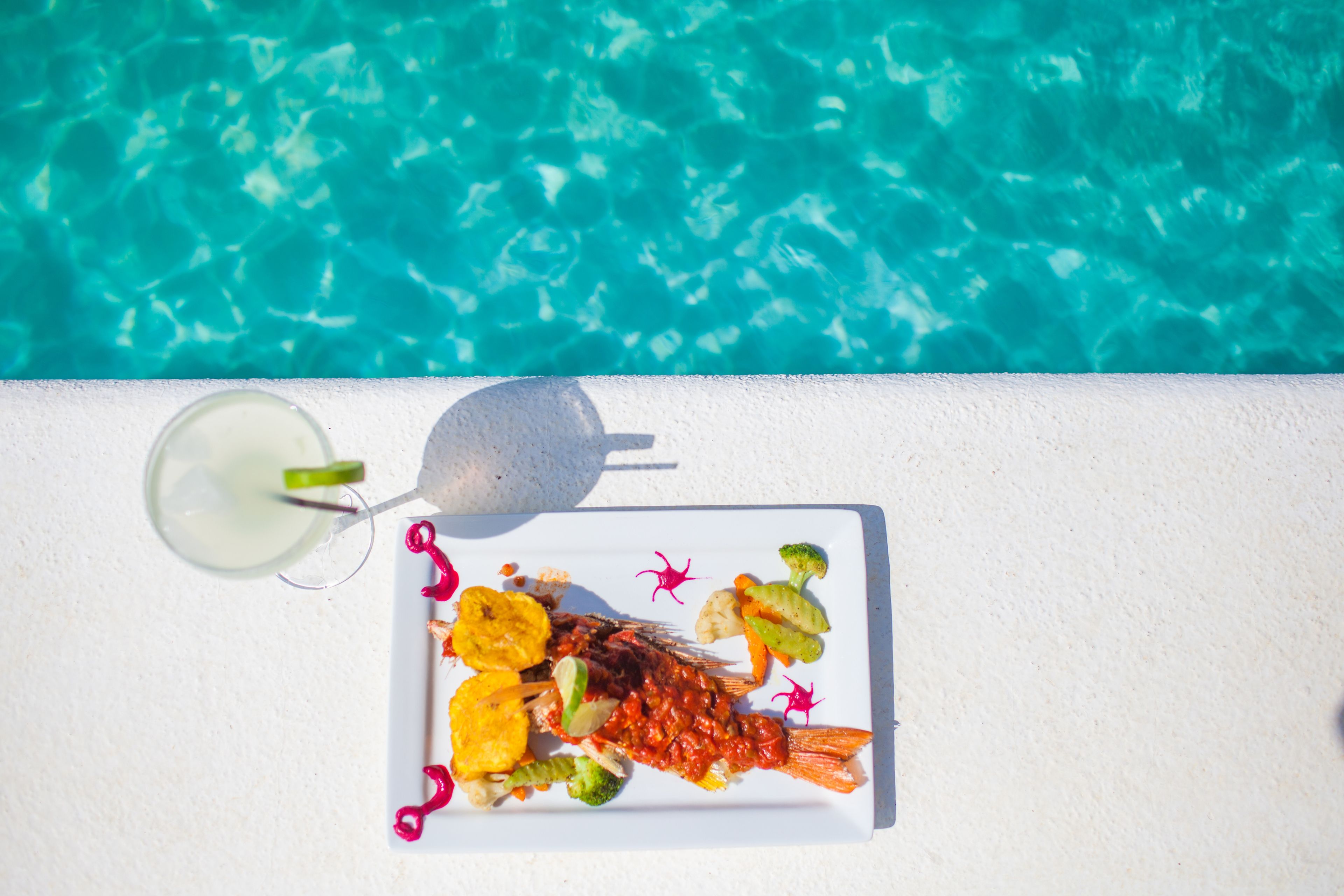 A white table with a plate of food and a drink by a pool.