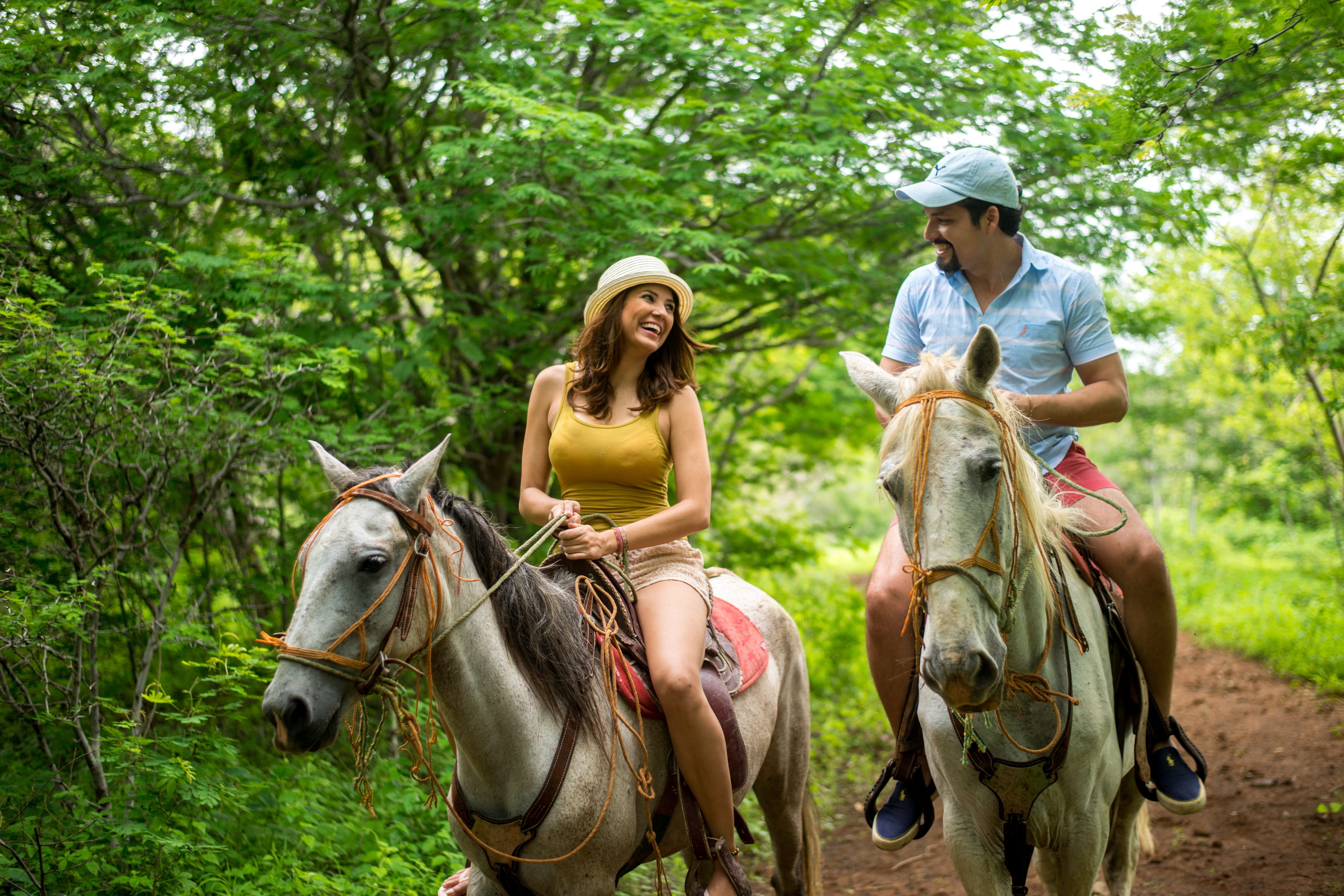 Two people riding horses on a dirt path surrounded by dense green foliage.