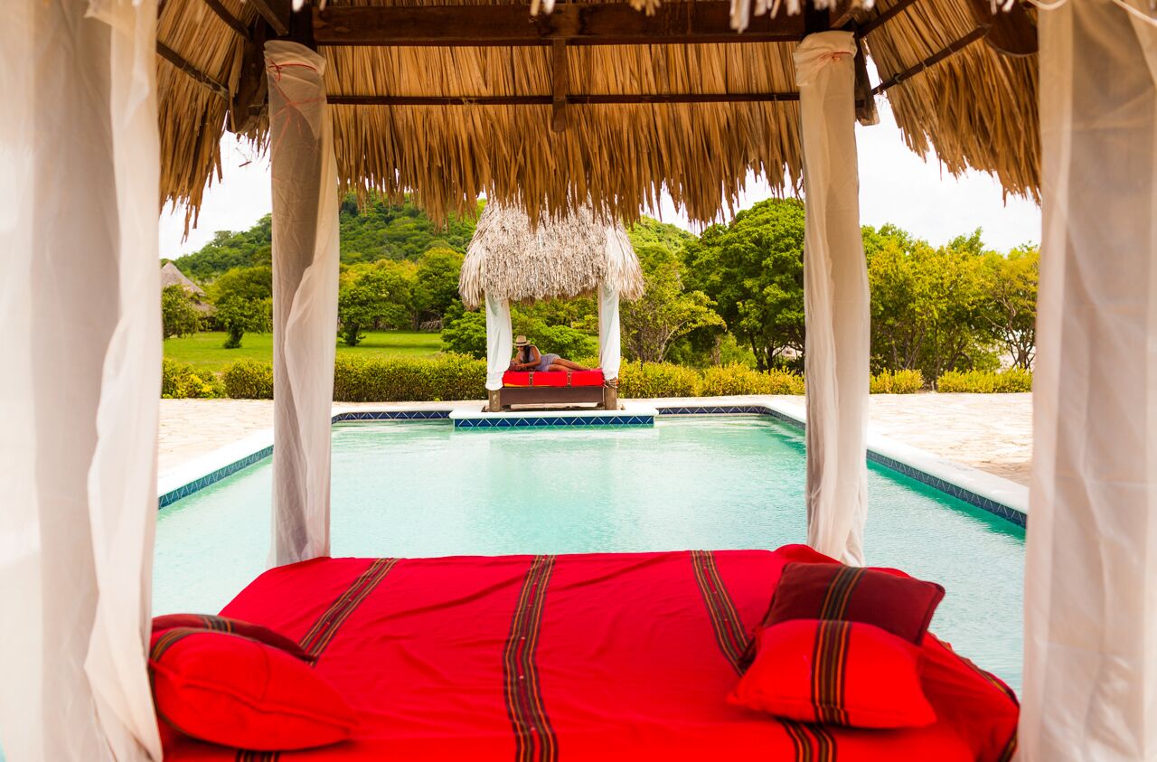 A bed with a red bedspread and pillows, a thatched roof structure, and a pool with a view of a lush green landscape.
