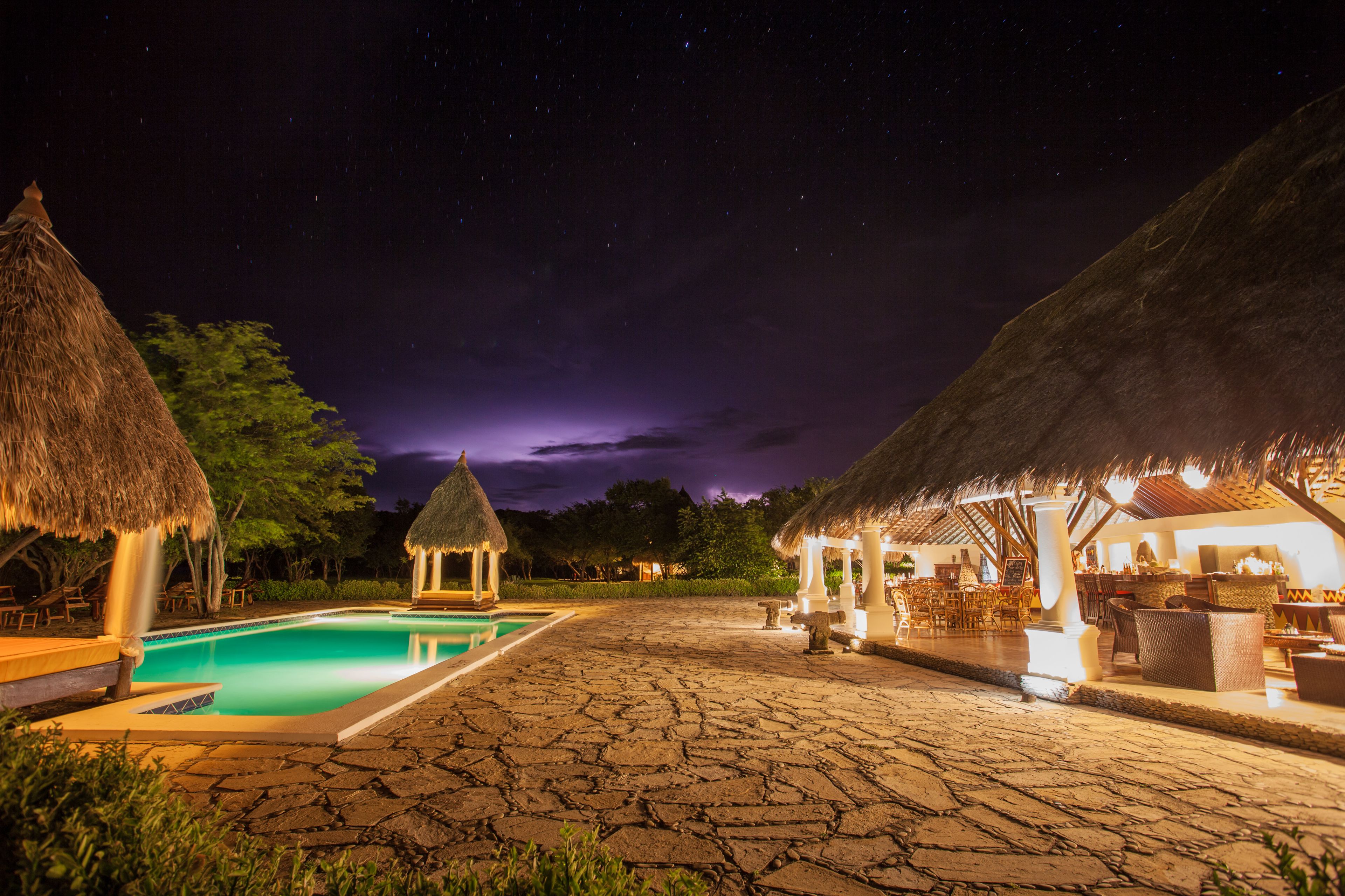 A nighttime resort with a swimming pool, thatched-roof structures, and a starlit sky.