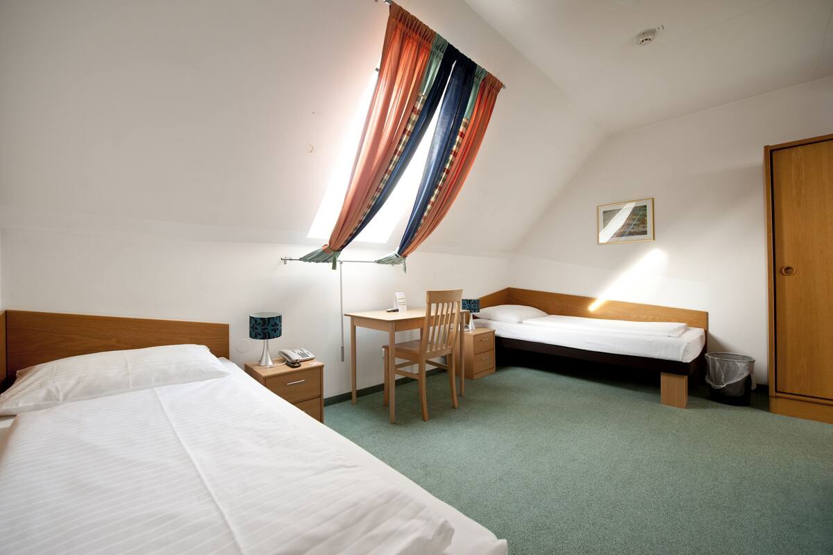 A hotel room with two beds, a desk, a chair, a small table, a trash bin, and a single window with curtains.