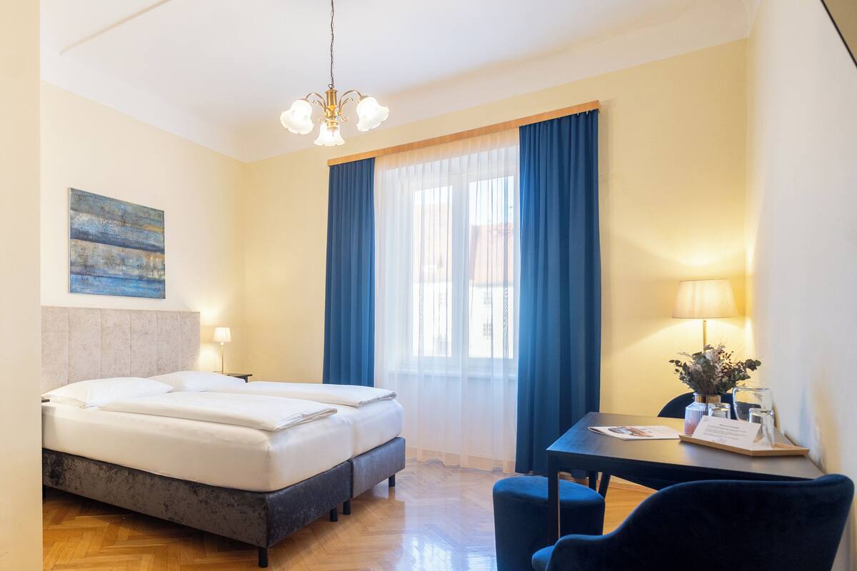 A hotel room with a bed, a desk with a chair, a lamp, and a window with curtains.
