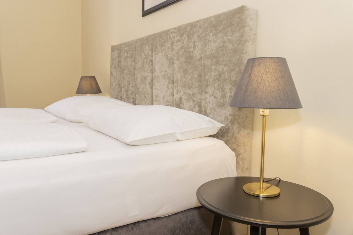 A neatly made bed with white linens and pillows, a round table with a lamp, and a textured headboard.
