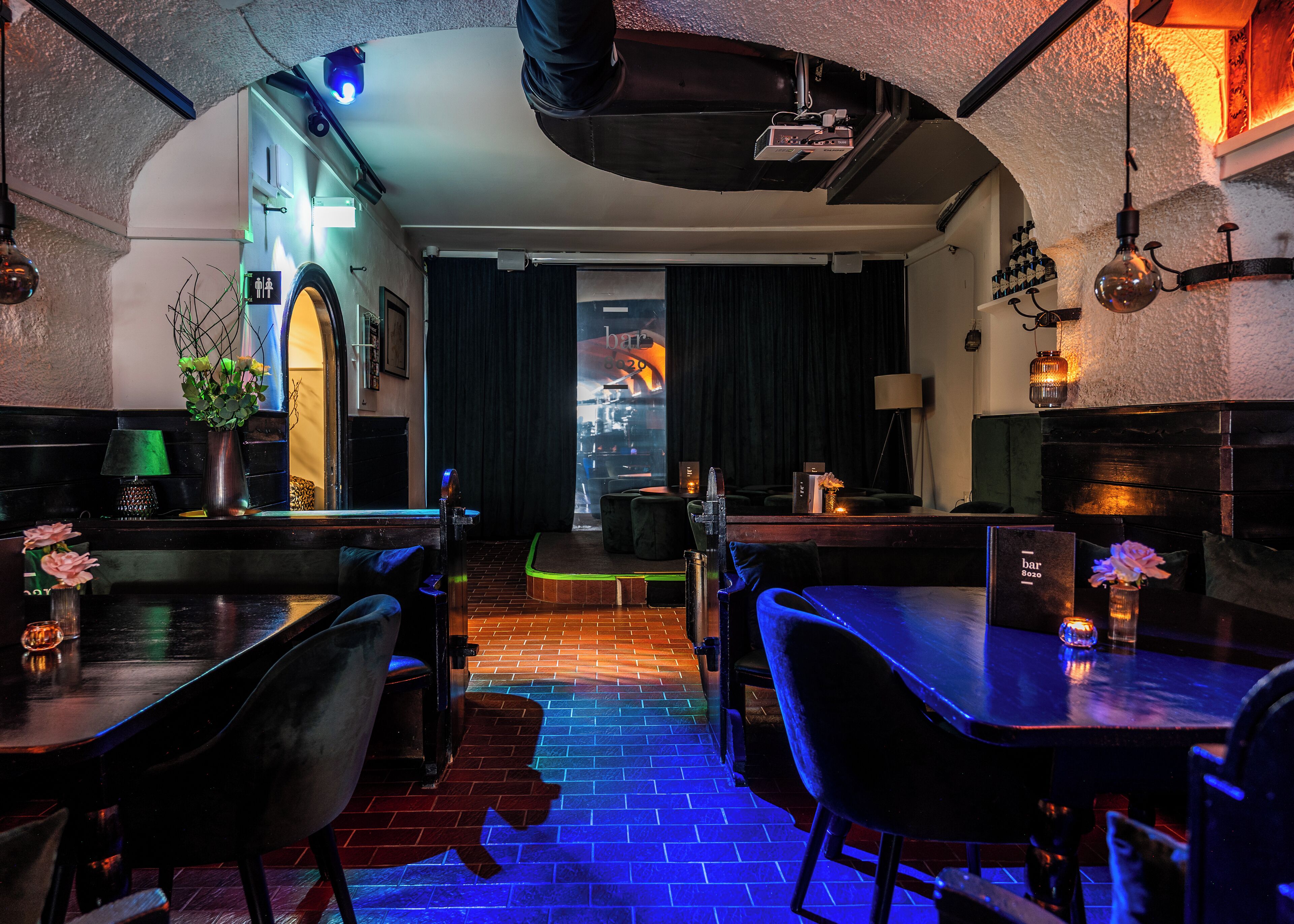 A bar with a brick floor and black furniture, a bar sign, and a projection screen in the background.