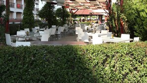 Un área de café al aire libre con mesas y sillas, cubierta por una estructura de madera con paneles blancos y rojos.