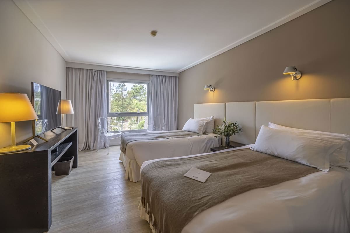 Habitación de hotel con dos camas, un escritorio con lámpara, televisión y ventana con cortinas.
