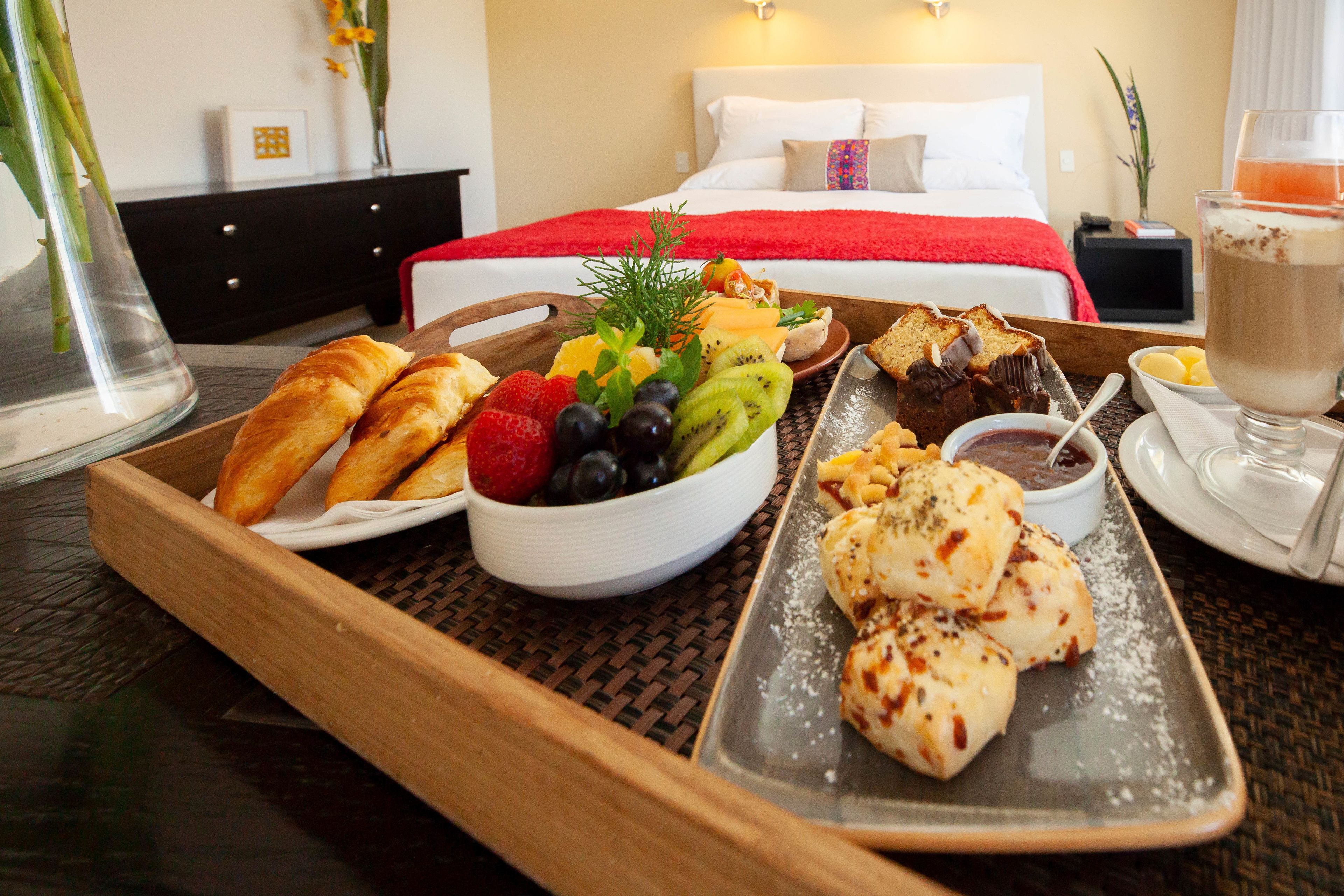 A breakfast tray with croissants, fruit, and pastries in a hotel room with a bed and nightstand in the background.