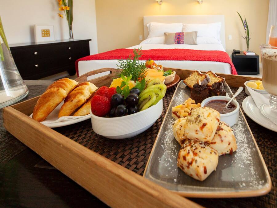 A breakfast tray with croissants, fruit, and pastries in a hotel room with a bed and nightstand in the background.