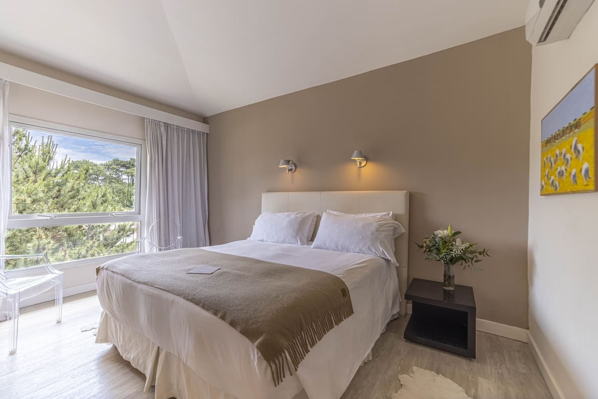 Un dormitorio con una cama grande, una mesita de noche con un florero, un cuadro en la pared y una ventana con cortinas.