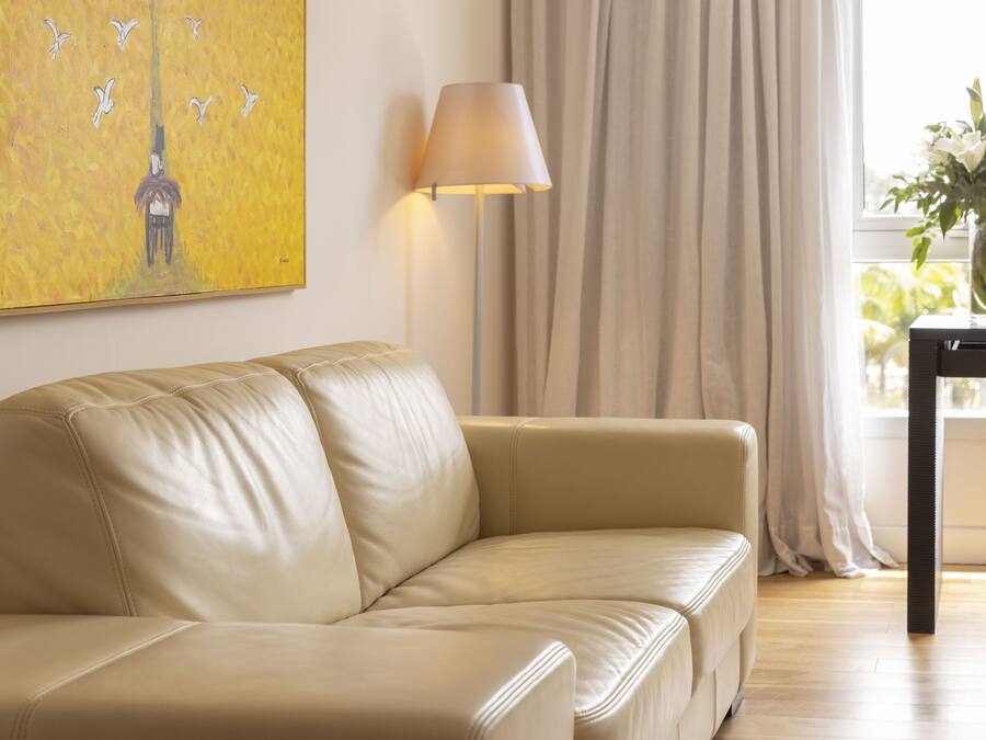 Un sofá de cuero color beige en una habitación bien iluminada, con un cuadro en la pared, una lámpara y una ventana con cortinas.