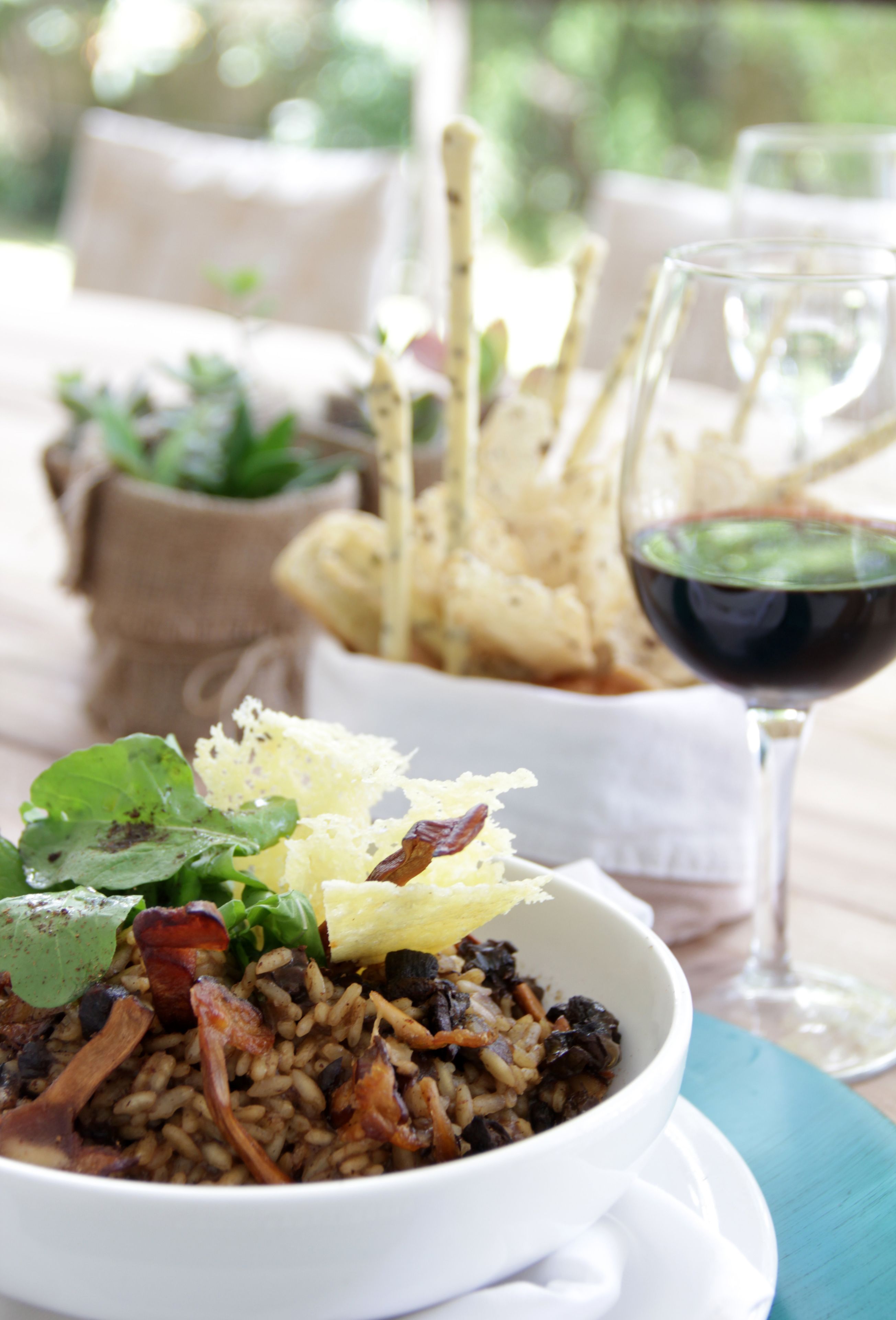 A bowl of rice with greens and bacon, a glass of red wine, and breadsticks on a table.
