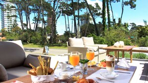 Une table de petit-déjeuner dressée avec des croissants, des pâtisseries, des fruits et des boissons, donnant sur un jardin luxuriant et de grands immeubles.
