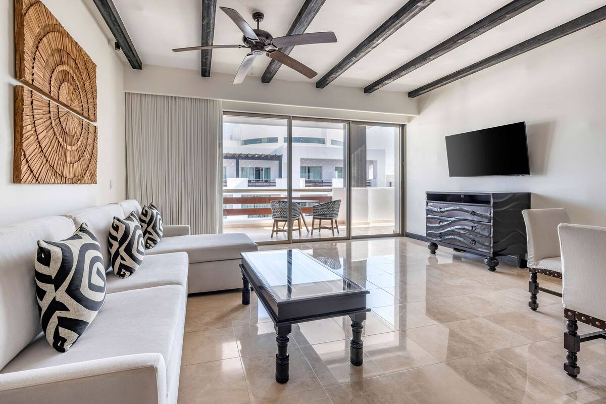 A modern living room with a glass coffee table, white sofa, patterned cushions, a ceiling fan, and a large window with a view of buildings.