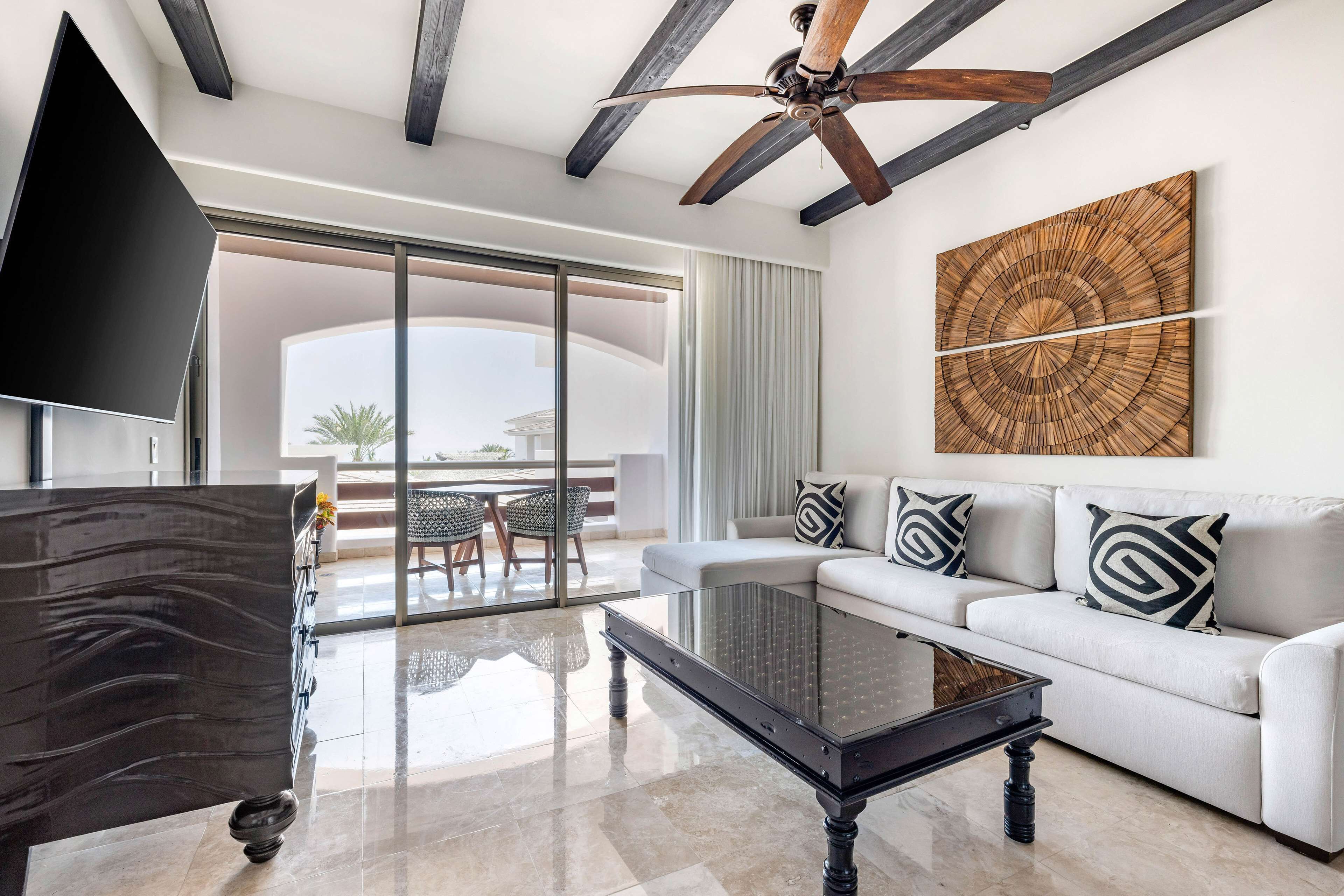 A modern living room with a glass coffee table, a sofa with patterned cushions, a ceiling fan, and a large wooden artwork on the wall.