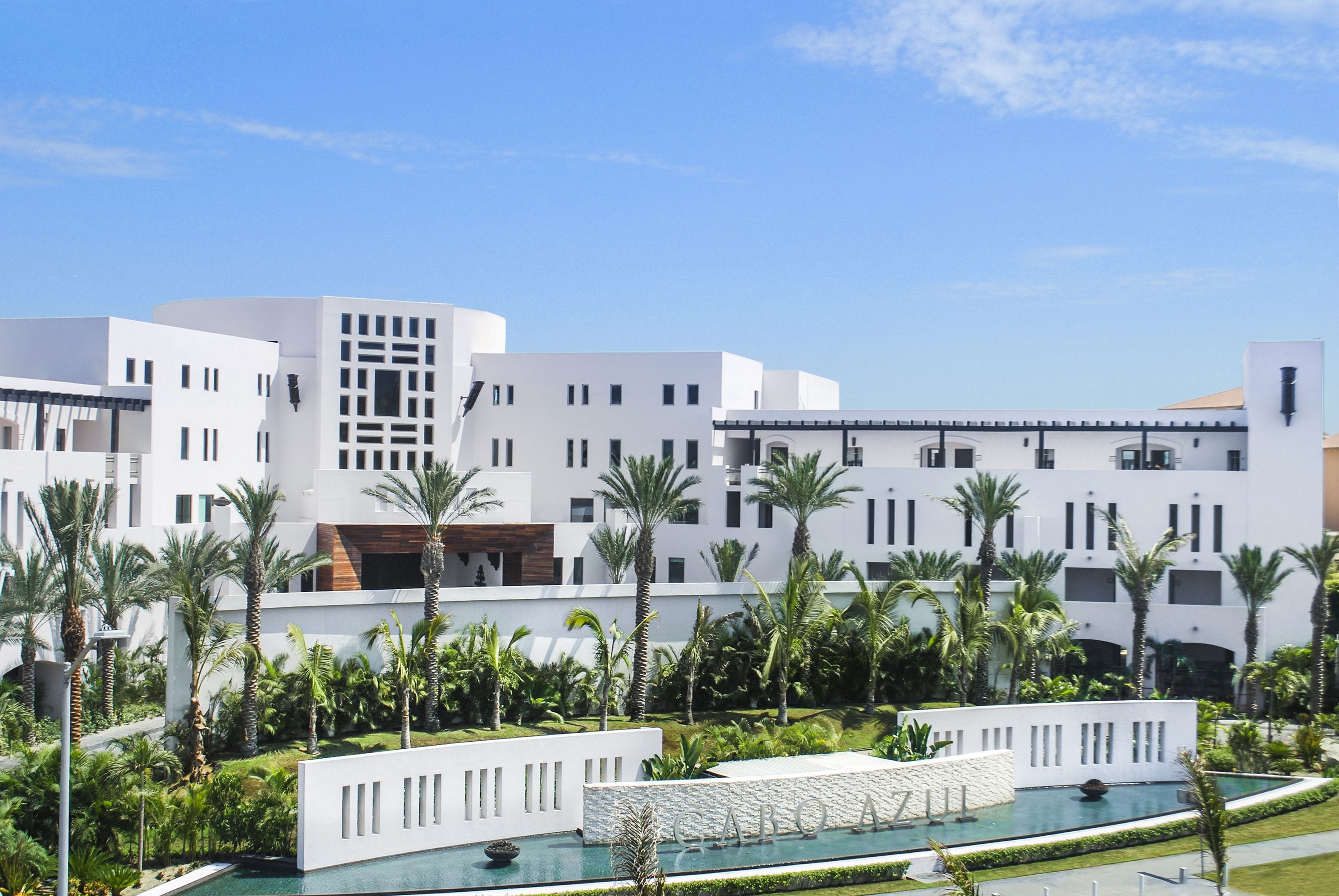 A modern hotel with white architecture, a pool, and palm trees.