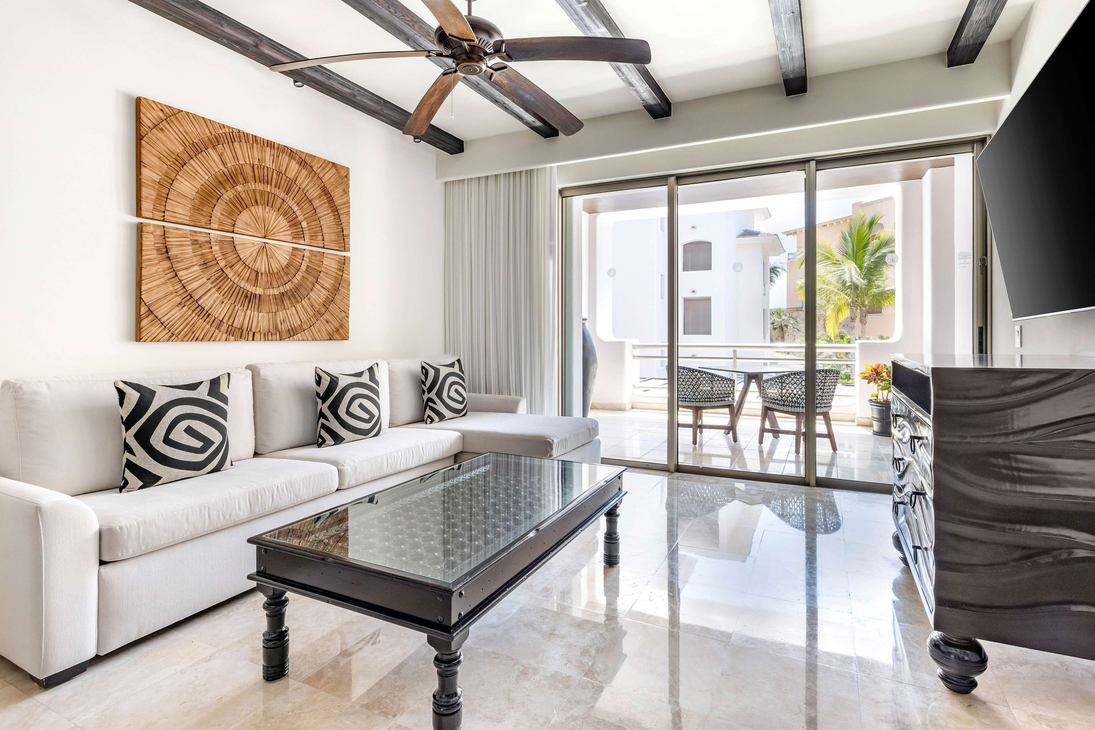 A modern living room with a white sofa, black and white patterned pillows, a glass coffee table, a ceiling fan, and a large window with a view of the outside.
