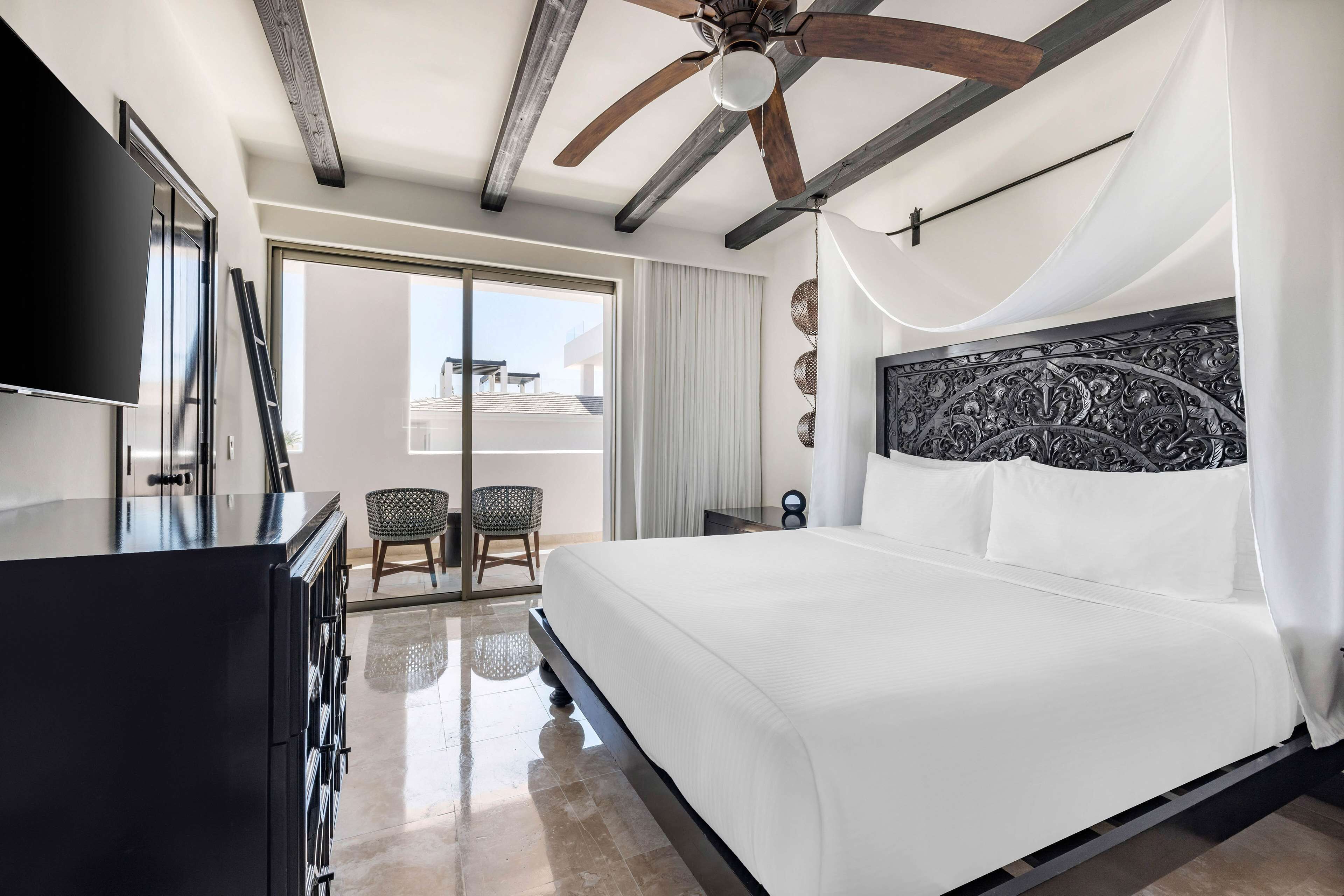Habitación de hotel moderna con una cama grande, ventilador de techo, televisor de pantalla plana y balcón con vistas.