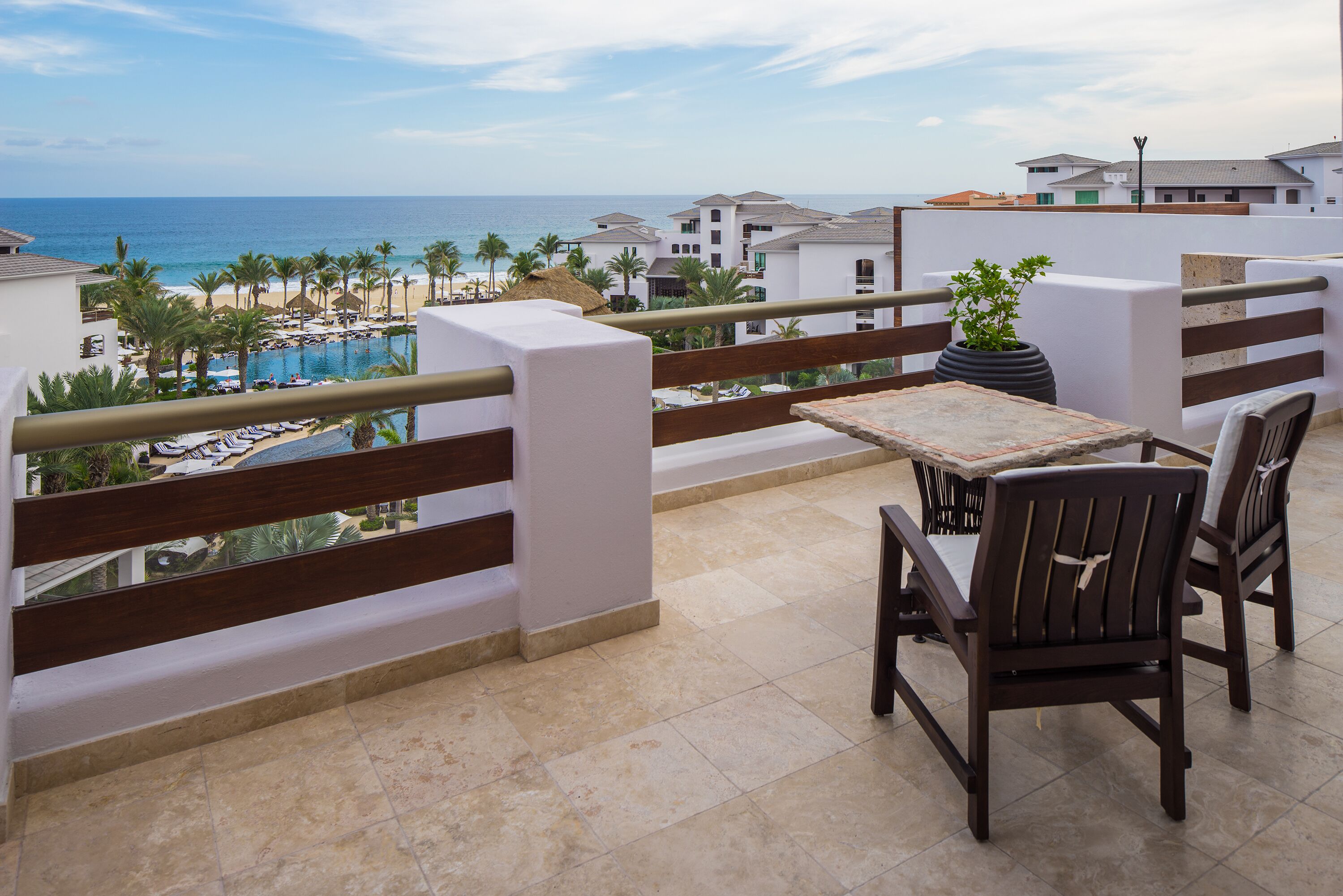 A balcony with a view of a beach and ocean, featuring a table and chairs, and a potted plant.