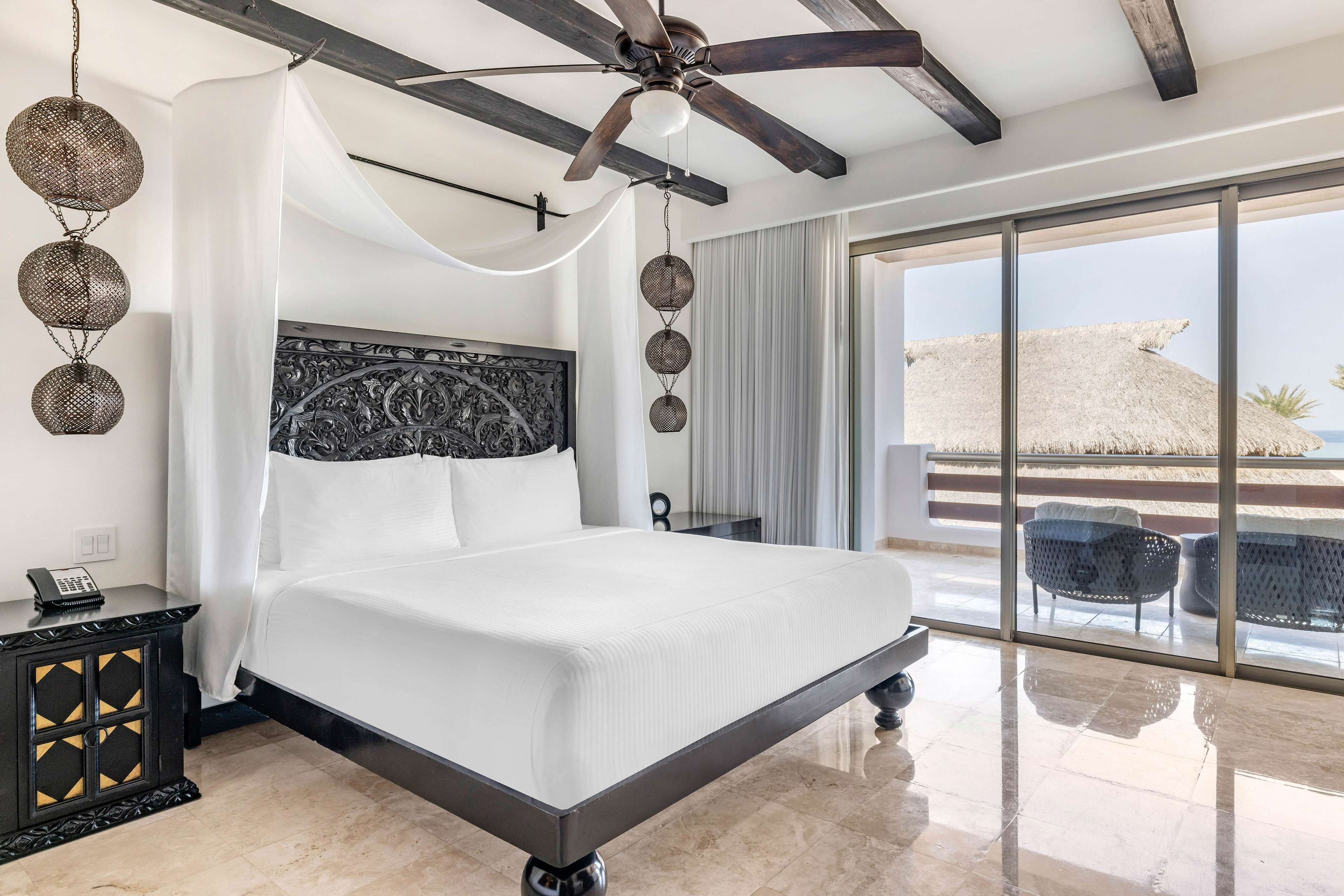 A modern hotel room with a large bed, a ceiling fan, and a view of the beach through the sliding glass door.