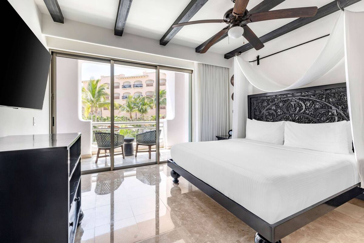 A hotel room with a large bed, a ceiling fan, a TV, and a balcony with a view of palm trees.