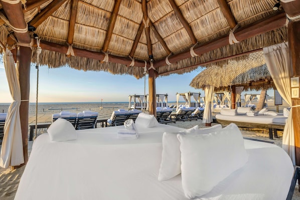 Habitación de hotel frente a la playa con techo de paja, ropa de cama blanca y vistas al mar.
