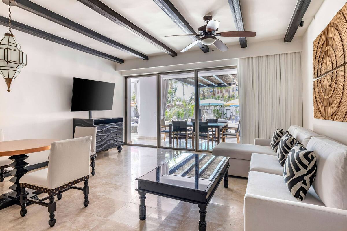 A modern living room with a glass coffee table, a sofa with patterned cushions, a flat-screen TV, and a ceiling fan.