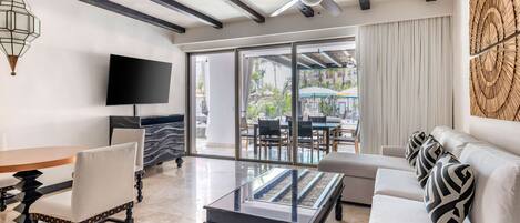 A modern living room with a glass coffee table, a sofa with patterned cushions, a flat-screen TV, and a ceiling fan.