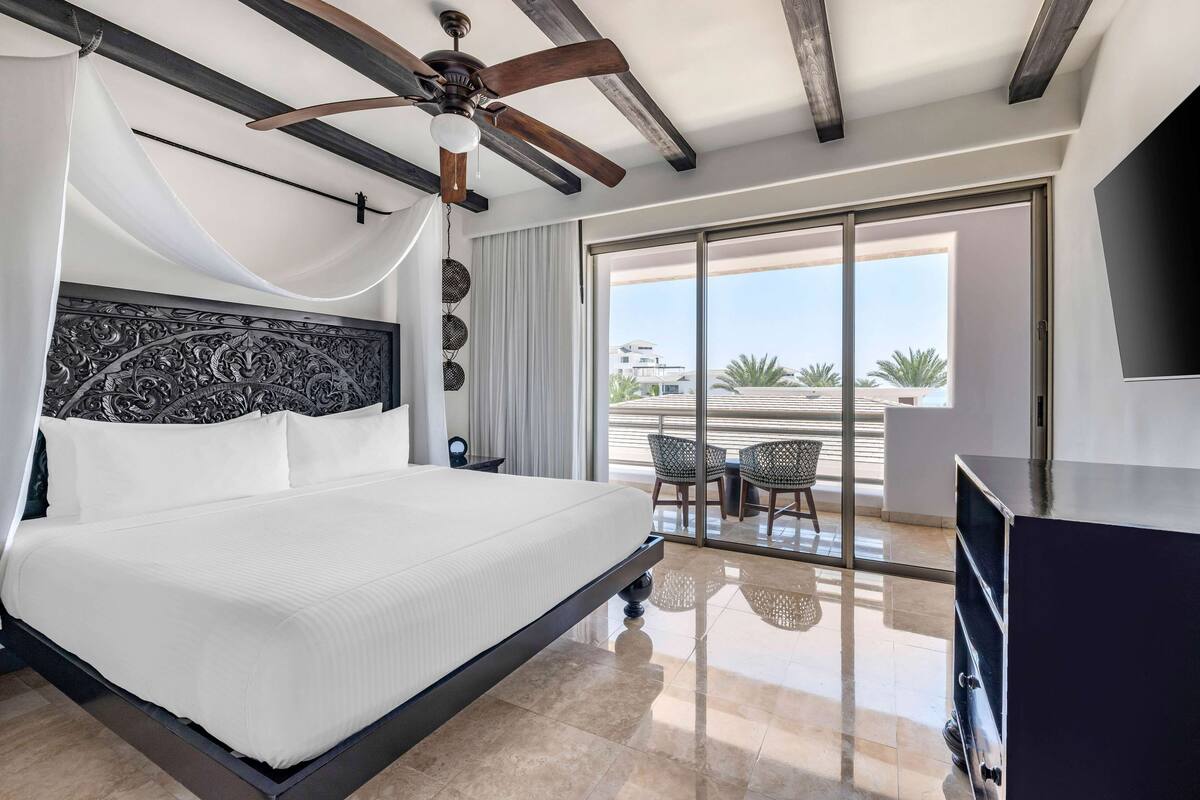 A hotel room with a large bed, a ceiling fan, a balcony with a table and chairs, and a TV.