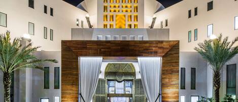 A modern hotel entrance with a central archway, large windows, and palm trees.