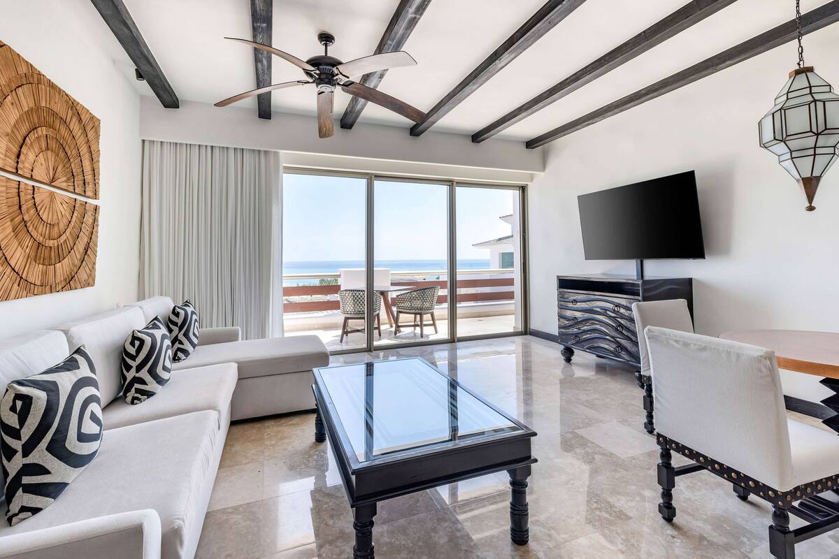 A modern living room with a glass coffee table, white sofas, and patterned cushions. A ceiling fan and a TV are mounted on the wall.