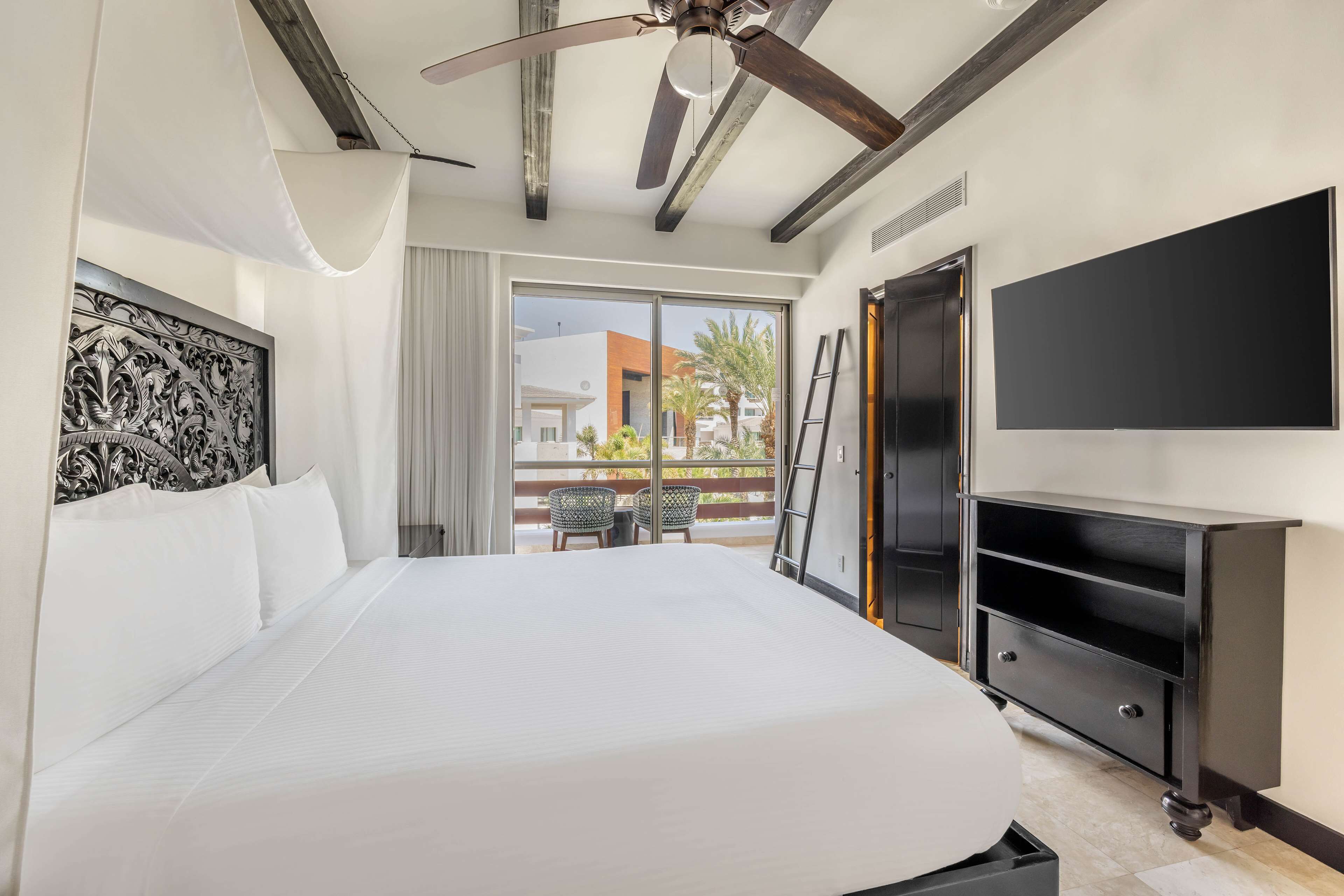 A hotel room with a large bed, a ceiling fan, a TV, and a balcony with outdoor seating.