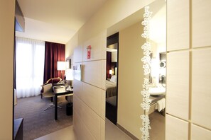 A hotel room with a desk, chair, and a bed. There is a bathroom visible through a mirror.