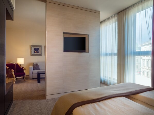 Quarto de hotel com cama, televisão na parede, sofá, poltrona e janela com cortinas.