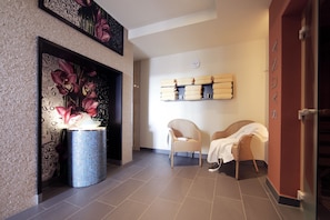 Un hall d’entrée moderne avec un sol carrelé, un mur décoratif orné d’une peinture florale, un vase bleu contenant des fleurs, deux fauteuils en osier et une étagère avec des serviettes.