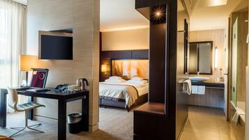 Habitación de hotel con cama, mesita de noche, lámpara, televisión y baño con lavamanos y espejo.