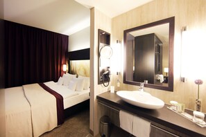 Habitación de hotel con cama, baño con lavamanos, espejo y artículos de aseo.