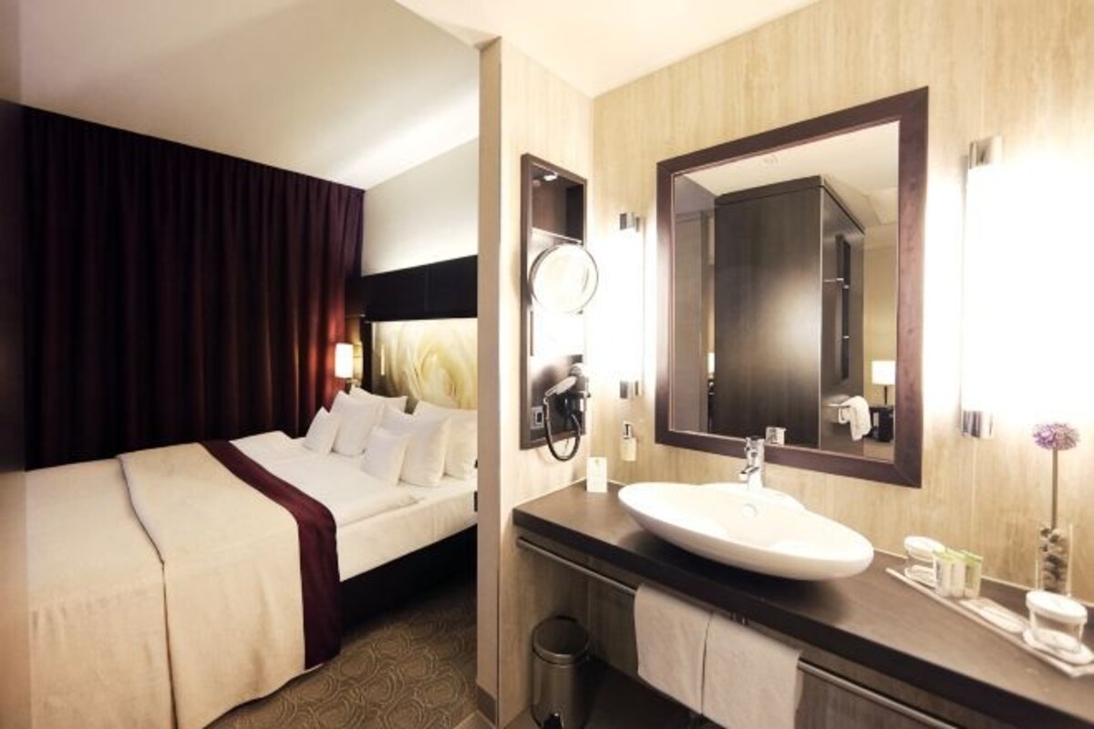 A hotel room with a bed, a large mirror, a sink, and a trash bin.