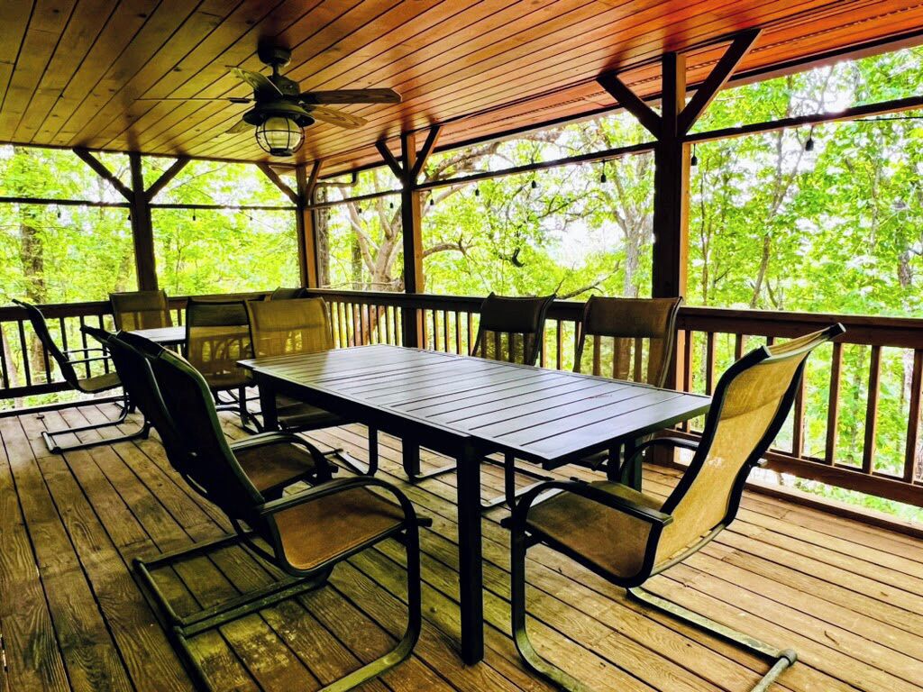 Un espacio de comedor al aire libre techado con mesa y sillas, piso de madera y vista a árboles a través de la barandilla.