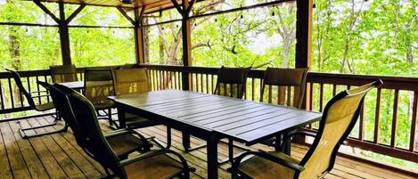 Un espacio de comedor al aire libre techado con mesa y sillas, piso de madera y vista a árboles a través de la barandilla.
