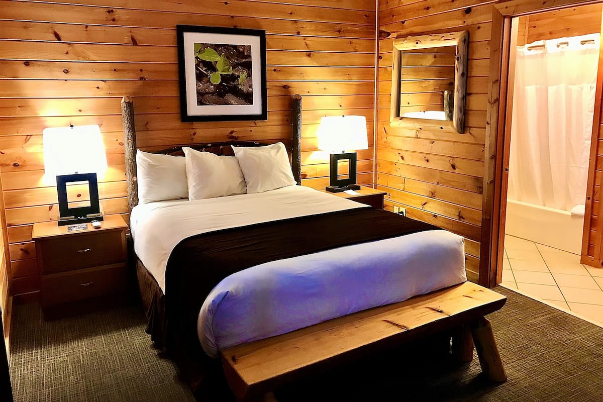 A wooden-paneled room with a bed, two bedside tables with lamps, a framed picture on the wall, and a mirror.