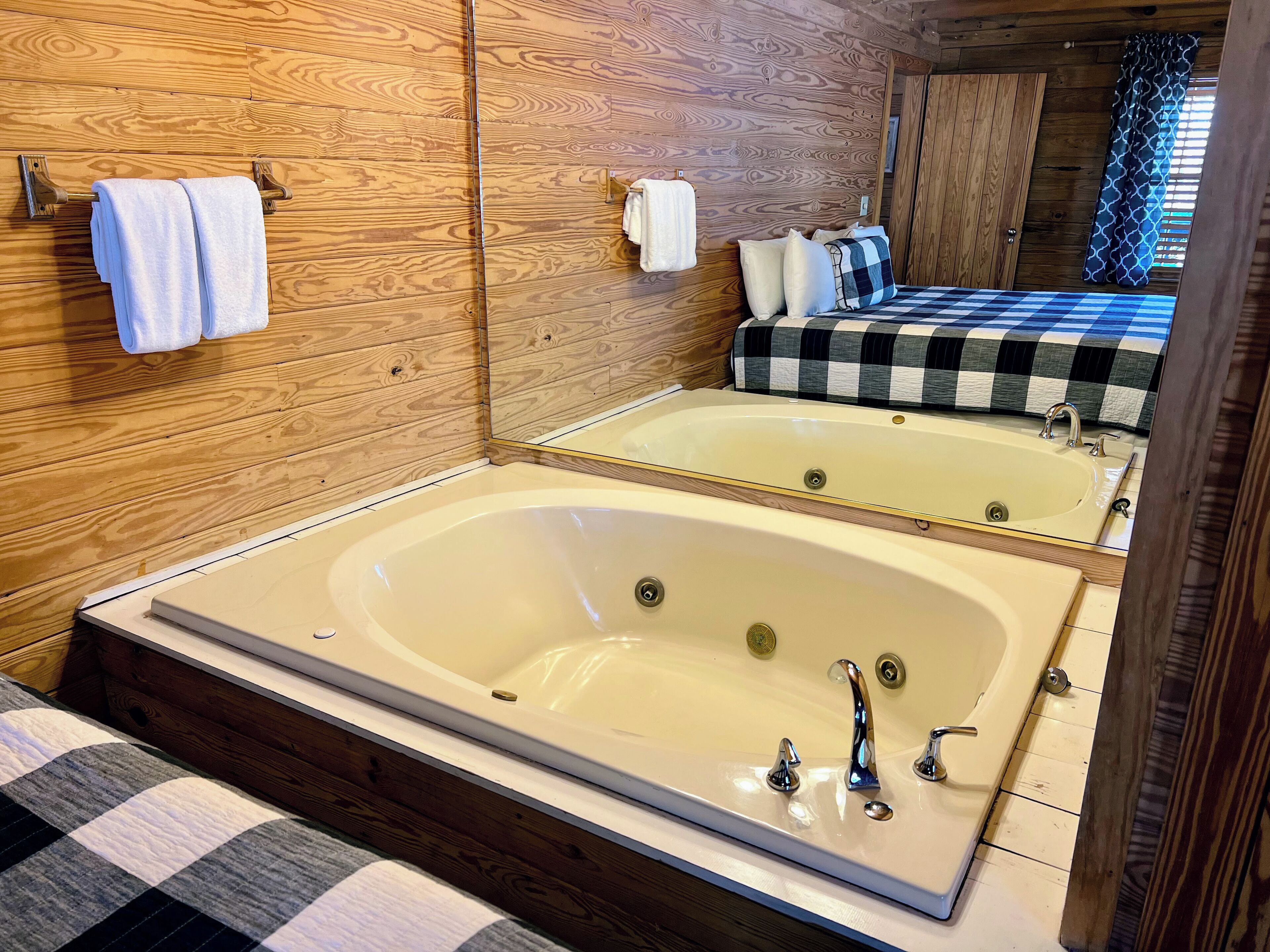 A wooden-paneled bathroom with a large bathtub, a mirror, and a bed with checkered bedding.