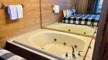A wooden-paneled bathroom with a large bathtub, a mirror, and a bed with checkered bedding.