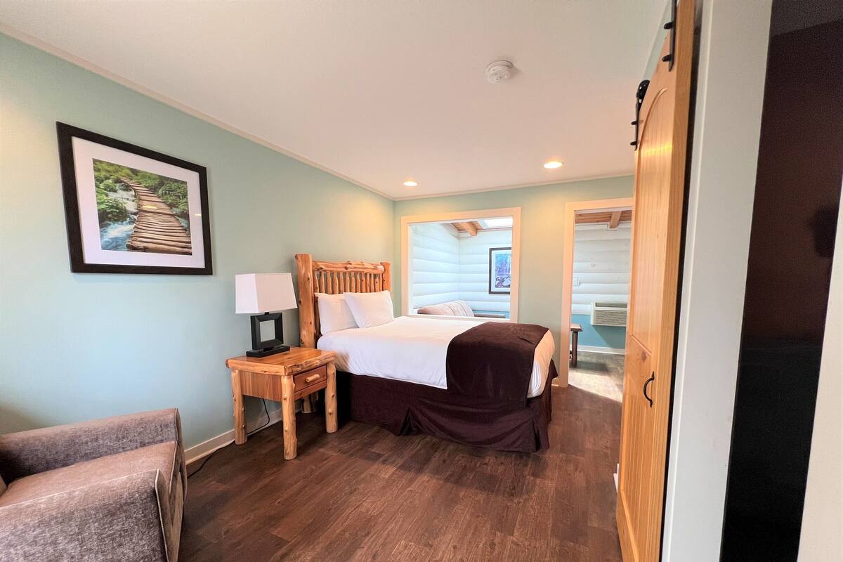 A hotel room with a wooden bed, a nightstand, a chair, and a framed picture on the wall.