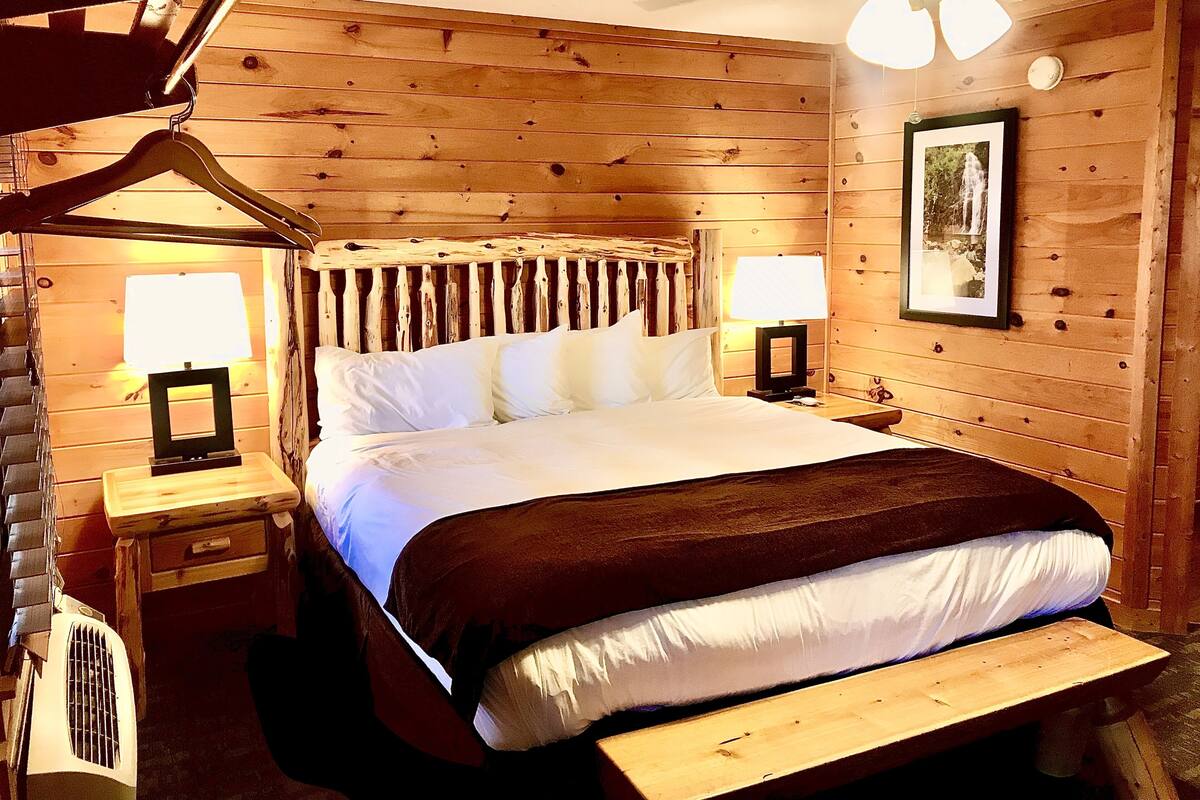 A wooden-paneled bedroom with a bed, two bedside tables with lamps, a framed picture on the wall, and a ceiling fan.