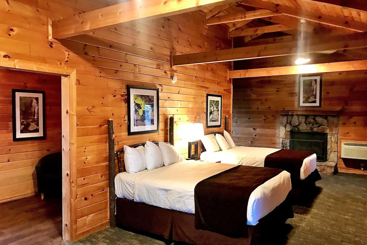 A wooden cabin-style room with two beds, a fireplace, and framed pictures on the walls.