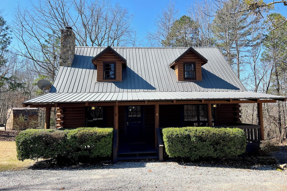 A log cabin with a metal roof, two dormer windows, a front door, and a gravel driveway.