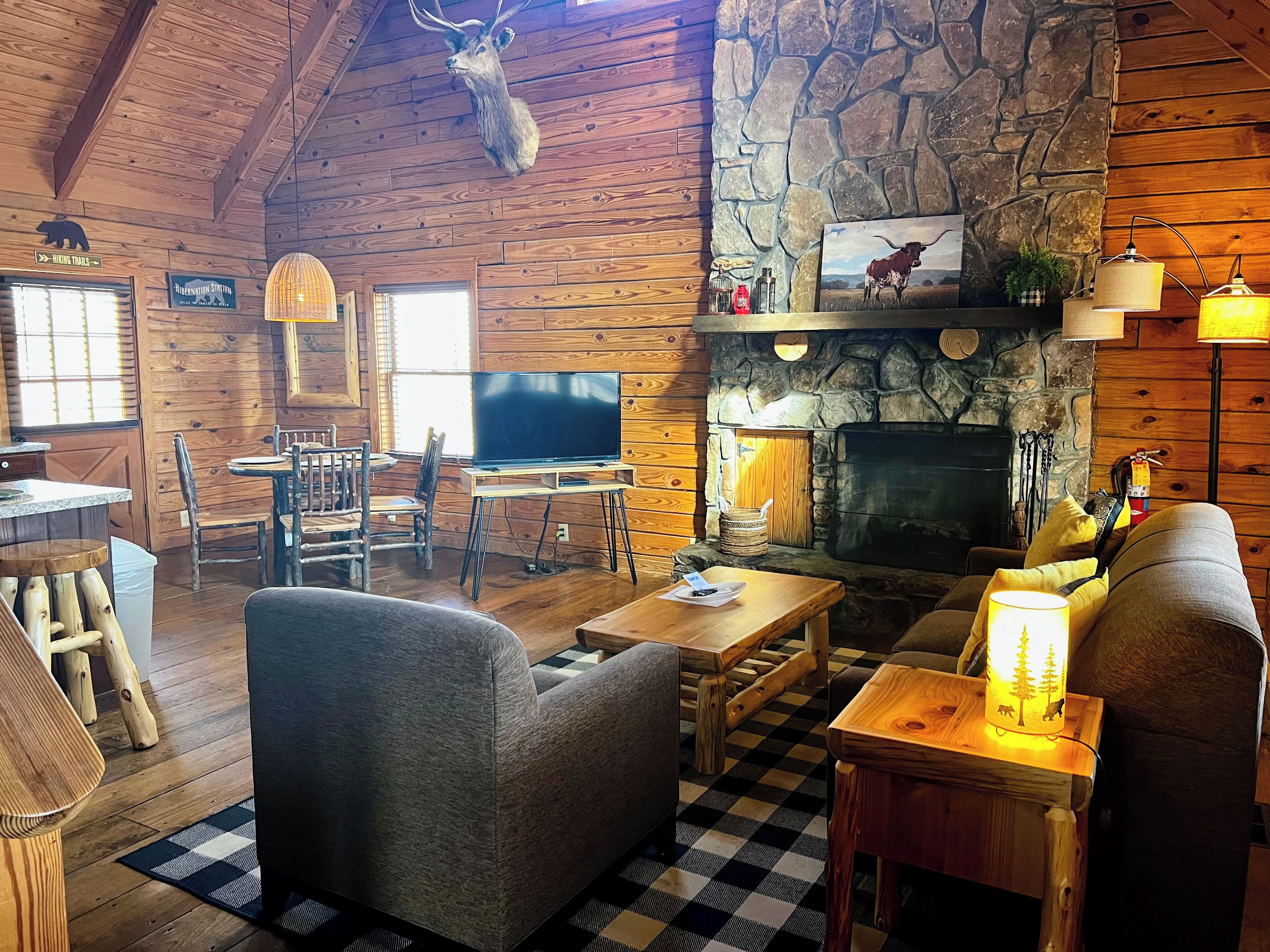 Interior de una cabaña de troncos con chimenea de piedra, muebles de madera y un venado disecado colgado.
