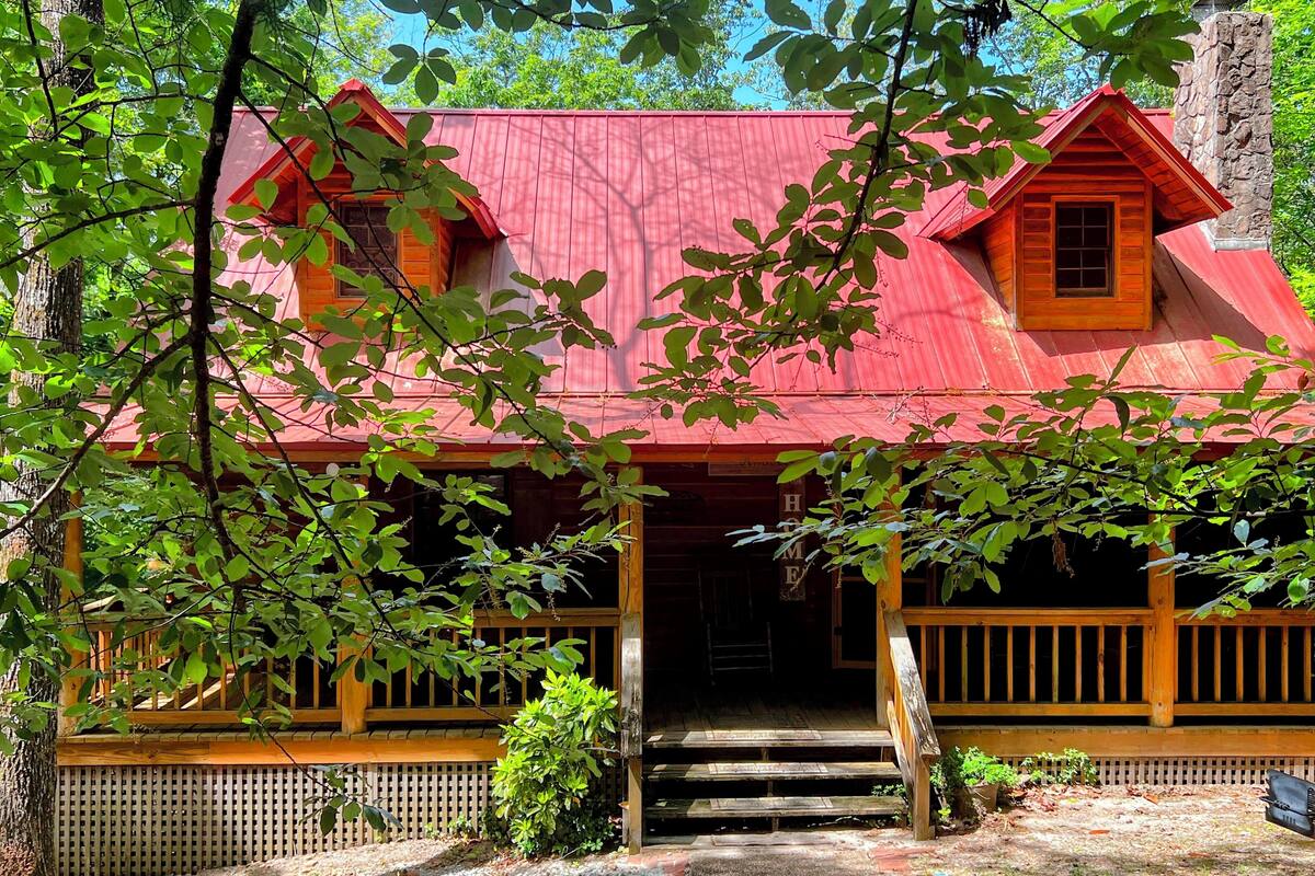 A rustic cabin with a red roof, wooden porch, and a staircase leading to the entrance.