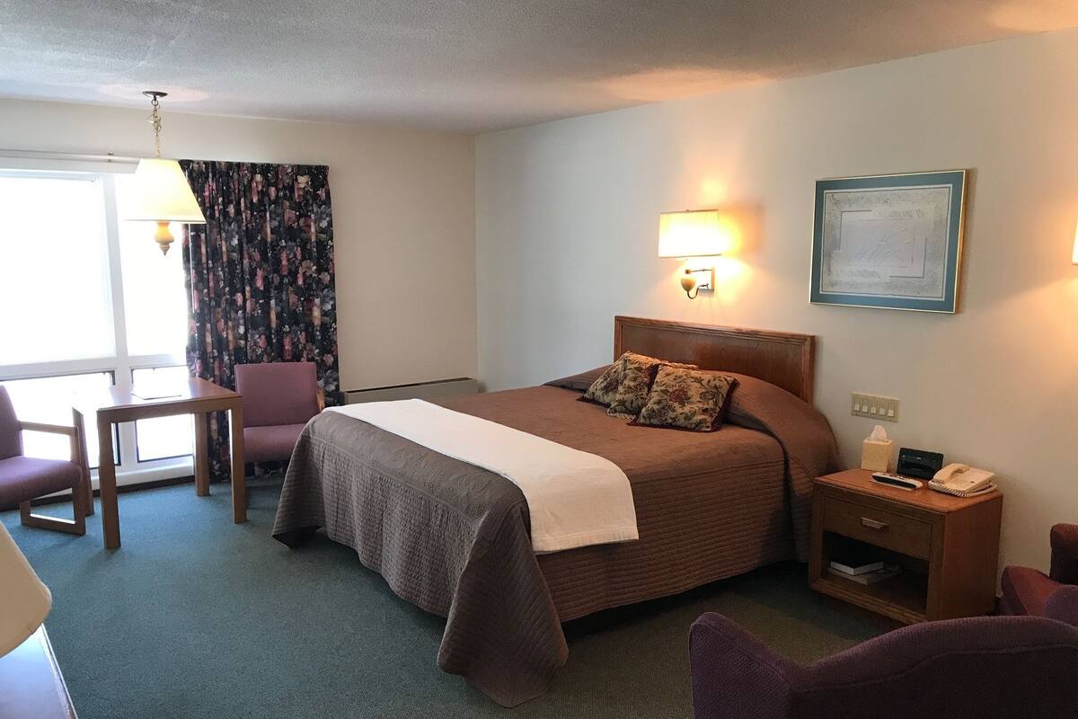 A hotel room with a bed, bedside table, lamp, chair, and a window with curtains.